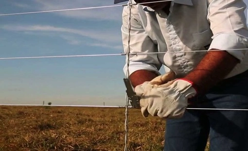 Imagem de um homem de camisa branca, com luva de segurança esticando o arame de cerca elétrica instalada em uma cerca em um pasto